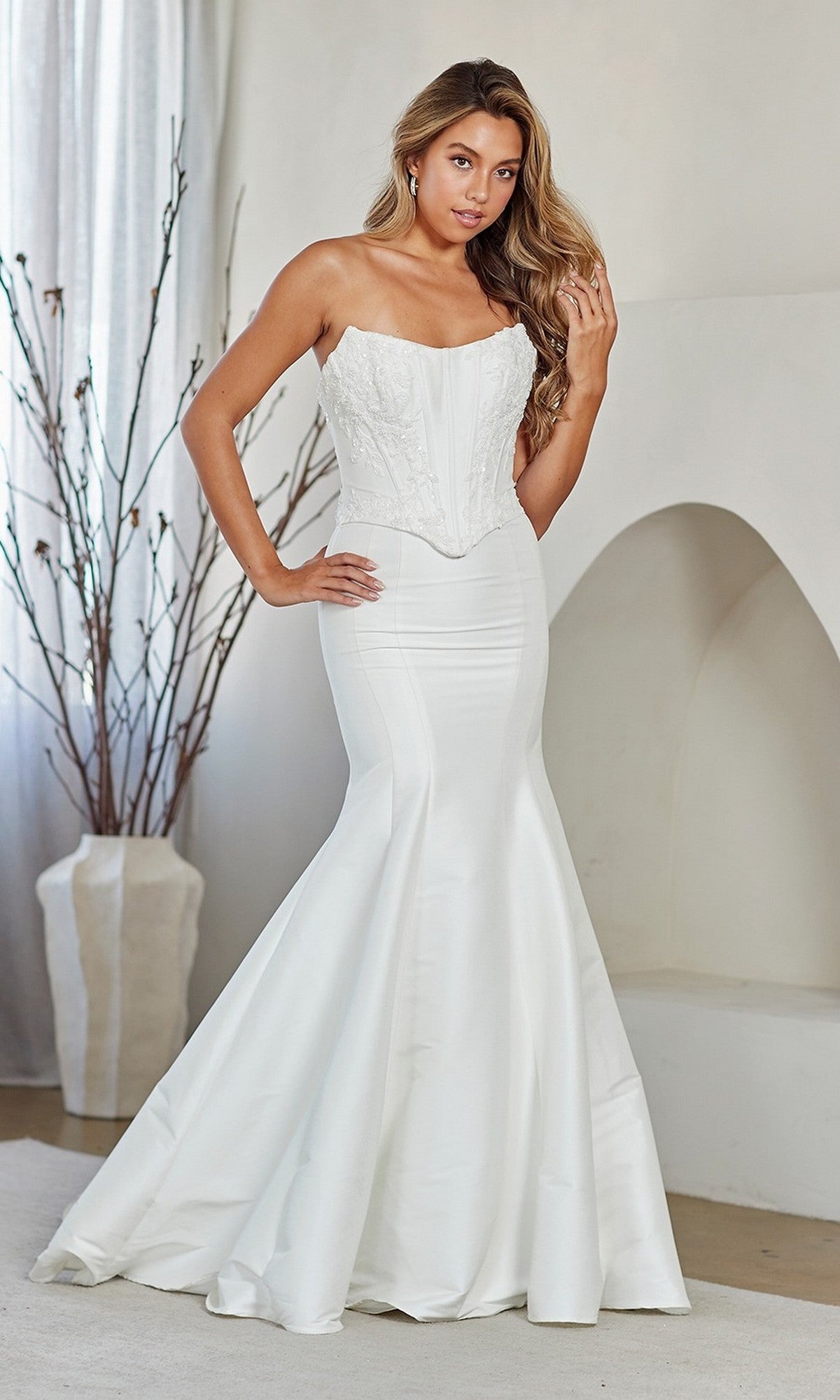 A woman stands indoors wearing the Amelia Couture 8080 long prom dress, featuring a strapless mermaid silhouette with a fitted embellished corset bodice and an elegant fit-and-flare skirt with minimal embroidery.